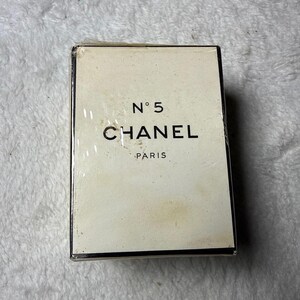 May include: A rectangular Chanel N&deg;5 perfume box with a cream-colored exterior and black lettering. The front of the box displays the text N&deg;5, CHANEL, and PARIS in a clean, sans-serif font. The packaging is encased in a clear plastic film wrap.