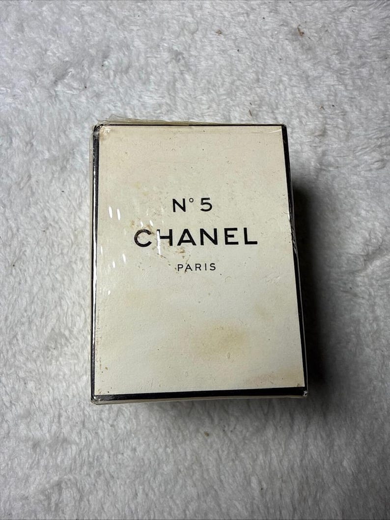 May include: A rectangular Chanel N&deg;5 perfume box with a cream-colored exterior and black lettering. The front of the box displays the text N&deg;5, CHANEL, and PARIS in a clean, sans-serif font. The packaging is encased in a clear plastic film wrap.
