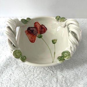 May include: A white ceramic bowl featuring a hand-painted red poppy flower with a green stem and bud. The bowl has two thick, twisted white handles on opposite sides, each accented with small, raised green spiral decorations at the base.