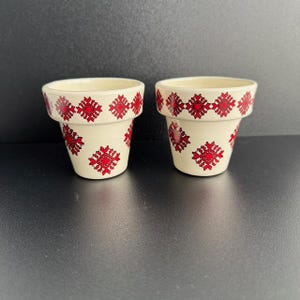 May include: Two white ceramic flower pots with red geometric patterns. The pots are empty and have a slightly rounded shape.