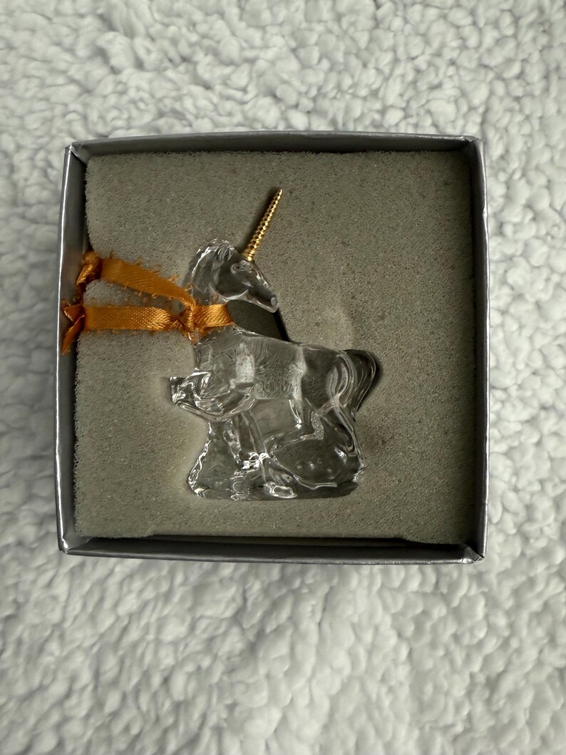 May include: A clear glass unicorn figurine with a gold horn, sitting in a grey box with a gold ribbon.
