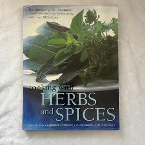 May include: A cookbook titled "Cooking with Herbs and Spices" with a green leafy herb arrangement on the cover. The book features over 200 recipes and is written by Andi Clevely, Katherine Richmond, Sallie Morris, and Lesley Mackley.