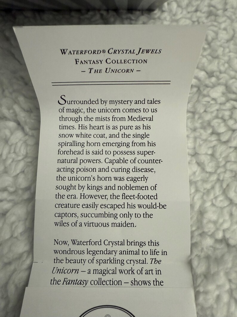 May include: A white card with black text describing the Waterford Crystal Fantasy Collection - The Unicorn. The text describes the unicorn as a mythical creature with magical powers and a symbol of purity. The card is surrounded by a white fuzzy background.