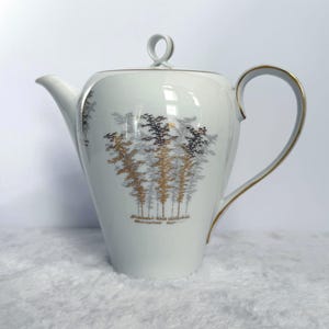 May include: White porcelain teapot with a lid and a curved handle. The teapot features a forest design in black, gray, and gold. The handle and rim are accented with gold trim. The spout is long and slender.