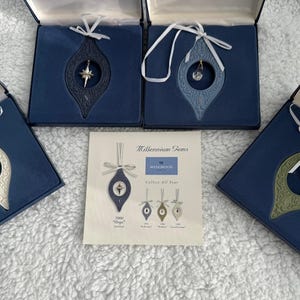 May include: Four blue boxes with white lids containing Wedgwood Millennium Gems ornaments. Each ornament is a teardrop shape with a different color and design. The boxes are on a white surface. The text on the card reads "Millennium Gems, Wedgwood, Collect All 12 Years, 2000 "Hope"