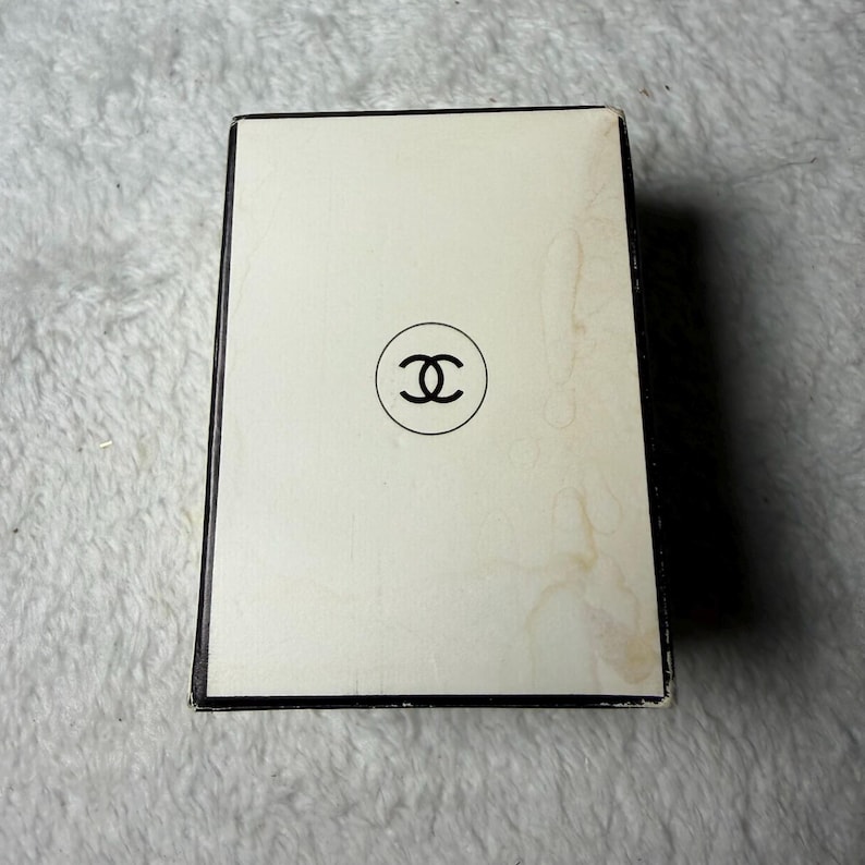 May include: A rectangular box with a cream-colored top surface featuring a black circular logo with two interlocking C letters in the center. The box has a black border and shows visible signs of discoloration and wear on the right side.