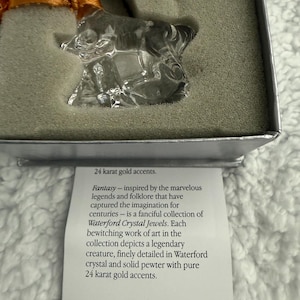 May include: A clear crystal unicorn ornament with 24 carat gold accents. The ornament is in a white box with a description of the ornament. The description reads: "Fantasy - inspired by the marvellous legends and folklore that have captured the imagination for centuries - is a fanciful collection of Waterford Crystal Jewels. Each bewitching work of art in the collection depicts a legendary creature, finely detailed in Waterford crystal and solid pewter with pure 24 carat gold accents."