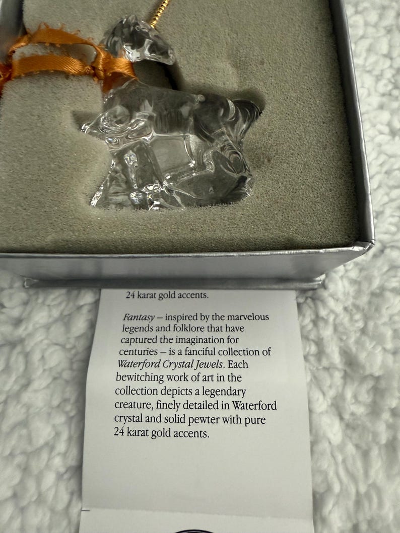 May include: A clear crystal unicorn ornament with 24 carat gold accents. The ornament is in a white box with a description of the ornament. The description reads: "Fantasy - inspired by the marvellous legends and folklore that have captured the imagination for centuries - is a fanciful collection of Waterford Crystal Jewels. Each bewitching work of art in the collection depicts a legendary creature, finely detailed in Waterford crystal and solid pewter with pure 24 carat gold accents."