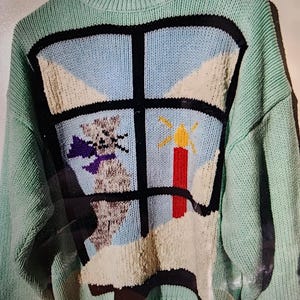 May include: A light green knit sweater with a whimsical design. The front features a window scene with a cat, sun, and snow. The window frame is black, and the sky is blue. The sweater has a ribbed neckline, cuffs, and hem.
