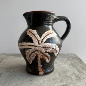 Puede incluir: Una pequeña jarra de cerámica negra con asa y pico. La jarra está decorada con un diseño de palmera blanca y marrón en la parte delantera. El borde de la jarra es marrón.