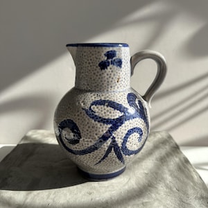Puede incluir: Una jarra de cerámica con cuerpo bulboso y asa. La jarra es blanquecina con textura moteada y está decorada con diseños florales y espirales azules. El borde y el asa también son azules. La jarra está sobre una superficie gris.