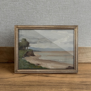 Puede incluir: Pintura de paisaje enmarcada que representa una escena costera con una playa de arena, acantilados y un cielo nublado. La obra de arte está encerrada en un marco rectangular de color dorado con un borde texturizado. Los colores de la pintura incluyen tonos de verde, beige y azul.