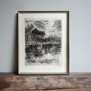 May include: Framed black and white etching of a serene landscape featuring a river, trees, and a full moon in the sky. The artwork is in a silver-toned frame, adding a classic touch to the piece. The print is signed.