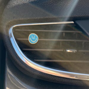 May include: A small, circular, light blue air freshener clip featuring a white smiley face icon attached to the horizontal slats of a black car dashboard air vent. The vent has a metallic silver trim and is positioned near the headlight controls.