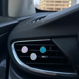 May include: Three circular car air vent clips featuring smiley face designs. The clips are colored light purple, bright blue, and white, each with a simple black line drawing of a smiling face, attached to the horizontal slats of a black car vent.