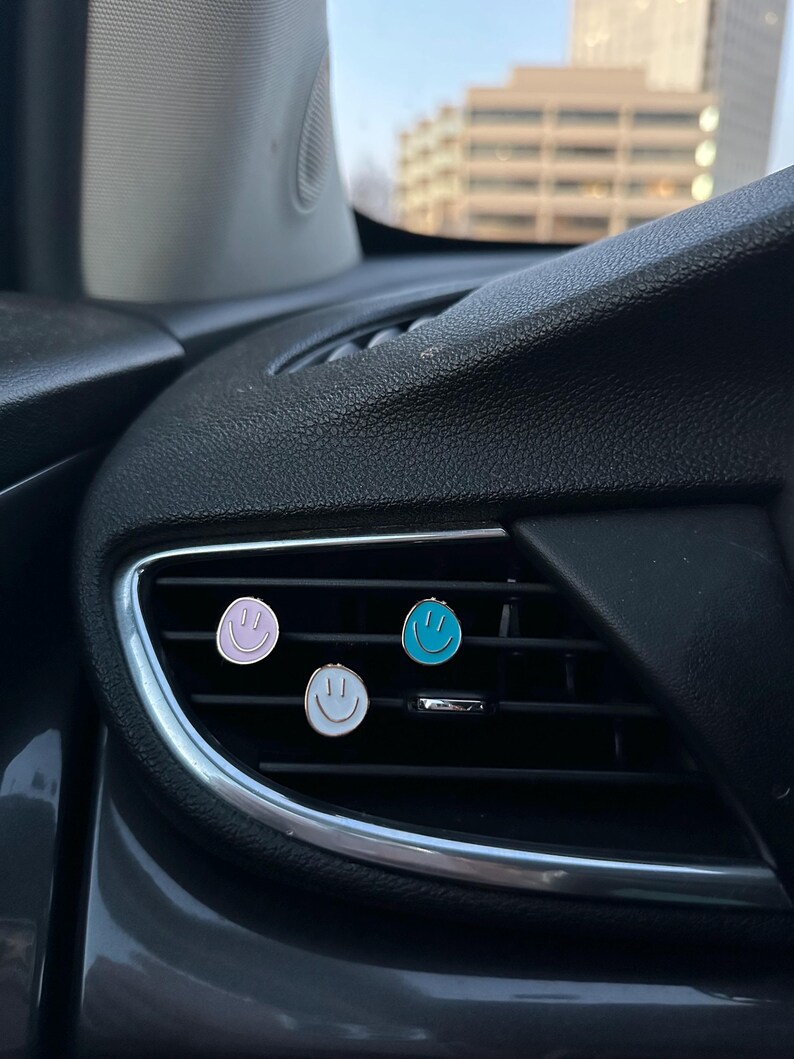 May include: Three circular car air vent clips featuring smiley face designs. The clips are colored light purple, bright blue, and white, each with a simple black line drawing of a smiling face, attached to the horizontal slats of a black car vent.