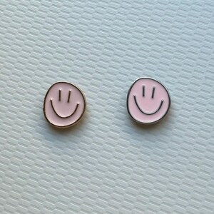 May include: Two circular enamel pins featuring a light pink smiley face design. The pin on the left has a gold-toned metal border, while the pin on the right features a silver-toned metal border. Both pins display two vertical eyes and a curved mouth.