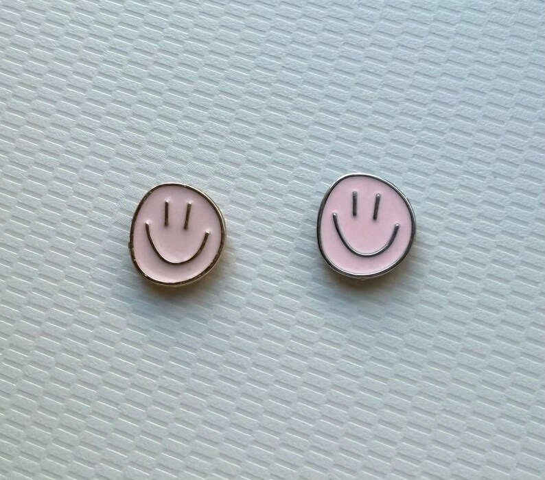 May include: Two circular enamel pins featuring a light pink smiley face design. The pin on the left has a gold-toned metal border, while the pin on the right features a silver-toned metal border. Both pins display two vertical eyes and a curved mouth.