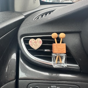 May include: A close-up of a car's interior with a wooden heart-shaped air freshener that says "Be Kind" and a wooden stick diffuser with a clear glass bottle.