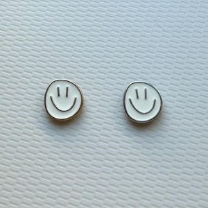 May include: Two circular stud earrings featuring a white enamel smiley face design. One earring has a gold-toned metal border, while the other earring features a silver-toned metal border. Both pieces display two vertical lines for eyes and a curved line for a mouth.
