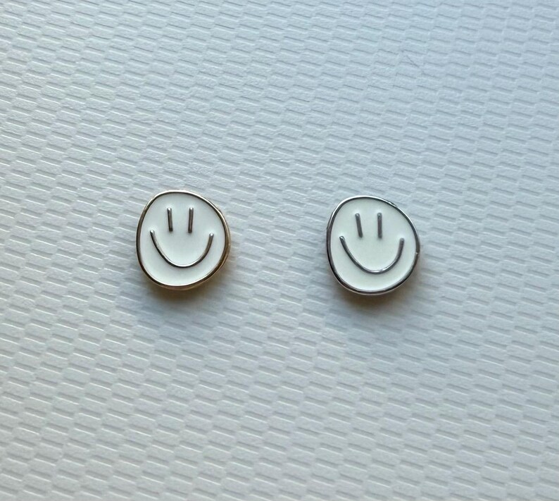 May include: Two circular stud earrings featuring a white enamel smiley face design. One earring has a gold-toned metal border, while the other earring features a silver-toned metal border. Both pieces display two vertical lines for eyes and a curved line for a mouth.