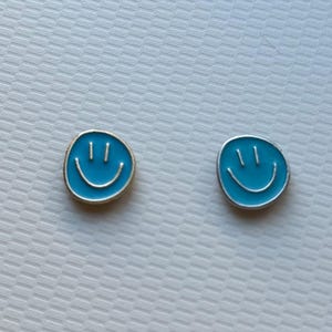 May include: A pair of circular stud earrings featuring a bright blue enamel finish with a silver-toned smiley face design. Each earring displays two vertical lines for eyes and a curved line for a mouth, set within a thin silver-colored metal border.