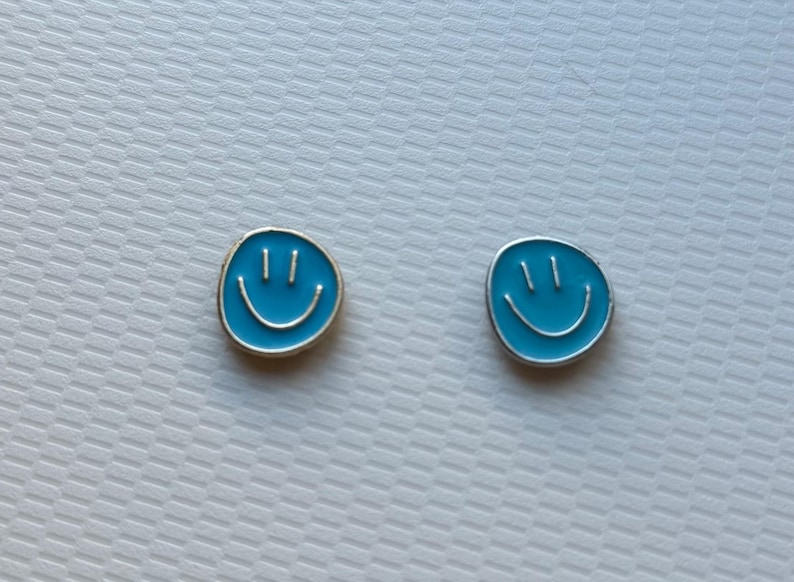 May include: A pair of circular stud earrings featuring a bright blue enamel finish with a silver-toned smiley face design. Each earring displays two vertical lines for eyes and a curved line for a mouth, set within a thin silver-colored metal border.