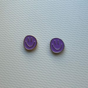 May include: A pair of circular stud earrings featuring a vibrant purple enamel finish. Each earring displays a simple, engraved smiley face design with two vertical lines for eyes and a curved line for a mouth, framed by a thin metallic border.