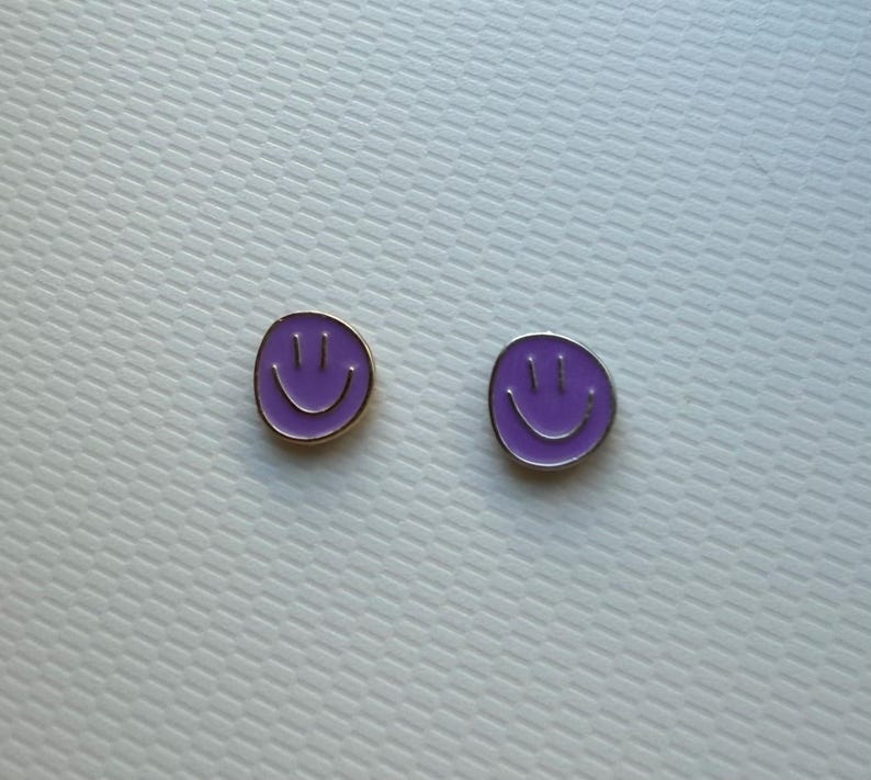 May include: A pair of circular stud earrings featuring a vibrant purple enamel finish. Each earring displays a simple, engraved smiley face design with two vertical lines for eyes and a curved line for a mouth, framed by a thin metallic border.