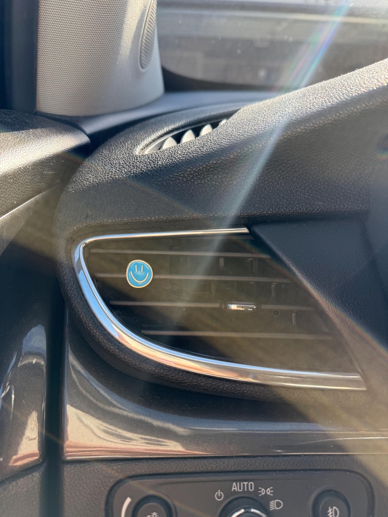 May include: A small, circular, light blue air freshener clip featuring a white smiley face icon attached to the horizontal slats of a black car dashboard air vent. The vent has a metallic silver trim and is positioned near the headlight controls.