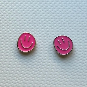 May include: Two small, circular metal pins featuring a vibrant pink enamel fill. Each pin displays a simple, etched smiley face design with two vertical lines for eyes and a curved line for a mouth, outlined in a polished silver-toned metal border.
