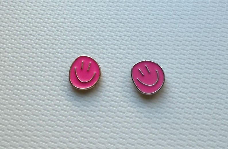 May include: Two small, circular metal pins featuring a vibrant pink enamel fill. Each pin displays a simple, etched smiley face design with two vertical lines for eyes and a curved line for a mouth, outlined in a polished silver-toned metal border.