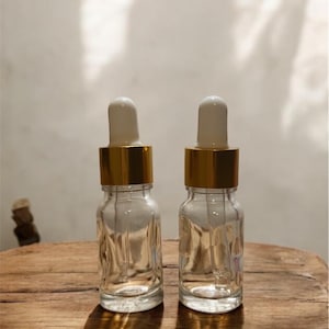 Peut inclure: Deux flacons compte-gouttes en verre transparent avec des colliers dorés et des dessus blancs sont posés sur une surface en bois. Les flacons ont un motif texturé. L'arrière-plan est de couleur blanc cassé neutre.