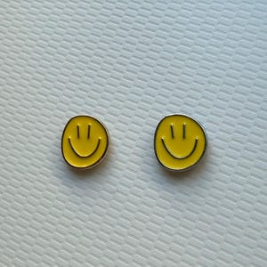 May include: A pair of circular stud earrings featuring a bright yellow enamel face with a black printed smiley design. Each earring has a simple, rounded shape with a smooth finish, showcasing two vertical eyes and a curved mouth in black.