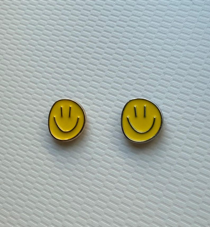 May include: A pair of circular stud earrings featuring a bright yellow enamel face with a black printed smiley design. Each earring has a simple, rounded shape with a smooth finish, showcasing two vertical eyes and a curved mouth in black.