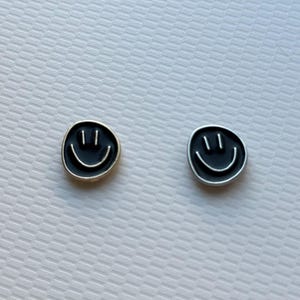 May include: A pair of circular stud earrings featuring a black enamel face with a silver-toned smiley face design. Each earring displays two vertical lines for eyes and a curved line for a mouth, set against a smooth, dark circular background.