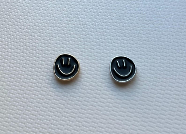 May include: A pair of circular stud earrings featuring a black enamel face with a silver-toned smiley face design. Each earring displays two vertical lines for eyes and a curved line for a mouth, set against a smooth, dark circular background.