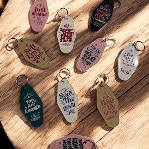 May include: A collection of motel-style keychains in various colors, each with inspirational phrases like "Just Breathe" and "Self Love Club." The keychains have metal rings and are arranged on a wooden surface.