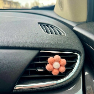 May include: A pink felt flower car air freshener with a white centre. The flower is attached to a car's air vent.