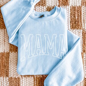 May include: Pale blue sweatshirt featuring the word "MAMA" in large white block letters. The sweatshirt has long sleeves and a crew neck. The garment is made of a soft material and is a light blue colour.