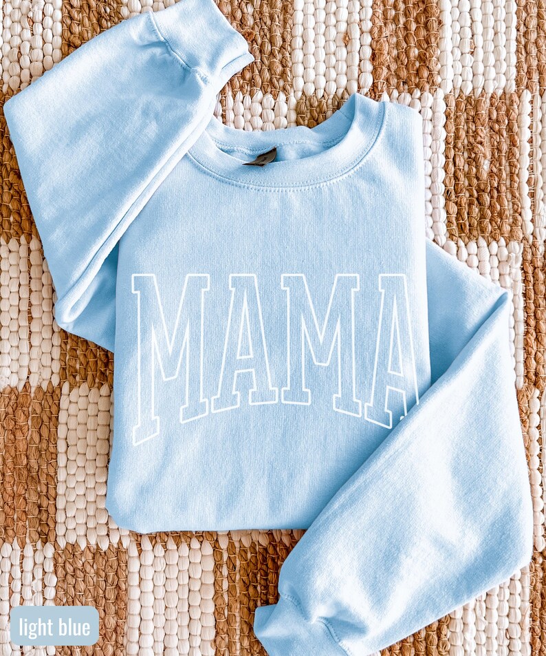 May include: Pale blue sweatshirt featuring the word "MAMA" in large white block letters. The sweatshirt has long sleeves and a crew neck. The garment is made of a soft material and is a light blue colour.