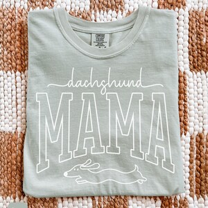 May include: A light green t-shirt with the words "dachshund MAMA" in white script and block letters. A white graphic of a dachshund dog is below the word "MAMA". The shirt is folded on a woven brown and white surface.