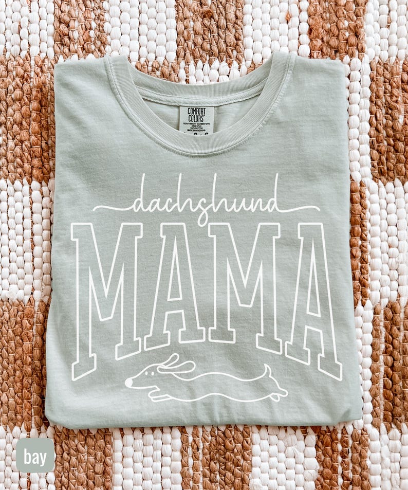 May include: A light green t-shirt with the words "dachshund MAMA" in white script and block letters. A white graphic of a dachshund dog is below the word "MAMA". The shirt is folded on a woven brown and white surface.