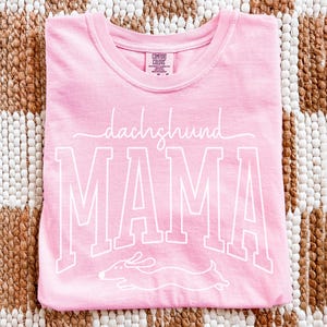 May include: A light pink t-shirt with the words "dachshund MAMA" in white script and block letters, along with a dachshund illustration. The shirt is folded and lies on a woven brown and beige surface.