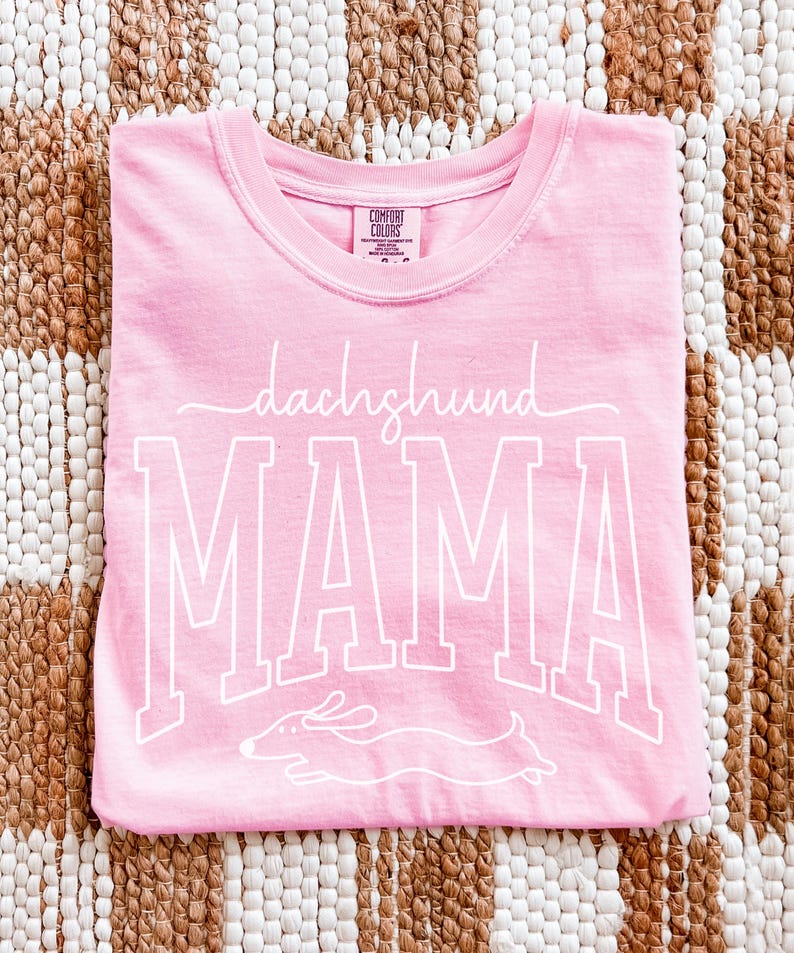 May include: A light pink t-shirt with the words "dachshund MAMA" in white script and block letters, along with a dachshund illustration. The shirt is folded and lies on a woven brown and beige surface.