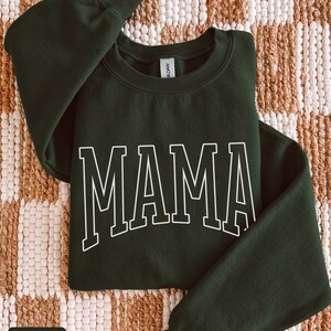 May include: A forest green sweatshirt featuring the word "MAMA" in white, block lettering. The garment is folded on a woven, beige and brown surface. The words "forest green" are in a small box in the lower left corner.