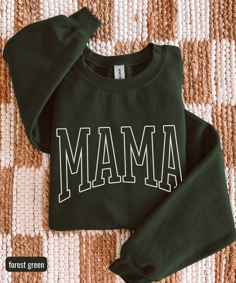 May include: A forest green sweatshirt featuring the word "MAMA" in white, block lettering. The garment is folded on a woven, beige and brown surface. The words "forest green" are in a small box in the lower left corner.