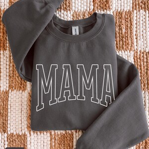 May include: A charcoal grey sweatshirt featuring the word "MAMA" in white, block lettering. The crew neck sweatshirt has long sleeves. The word "charcoal" is in a small box in the lower left corner.