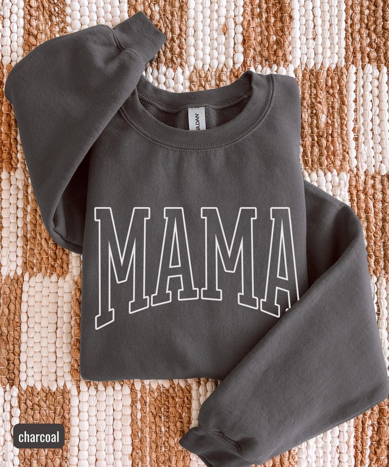 May include: A charcoal grey sweatshirt featuring the word "MAMA" in white, block lettering. The crew neck sweatshirt has long sleeves. The word "charcoal" is in a small box in the lower left corner.