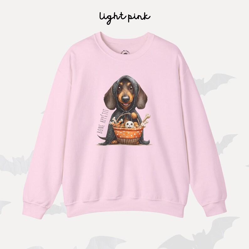 May include: Pale pink sweatshirt with a dachshund illustration. The dog wears a cloak and holds a basket of bones and a small dog. The words "BONE APPETIT" are printed on the left side of the sweatshirt.